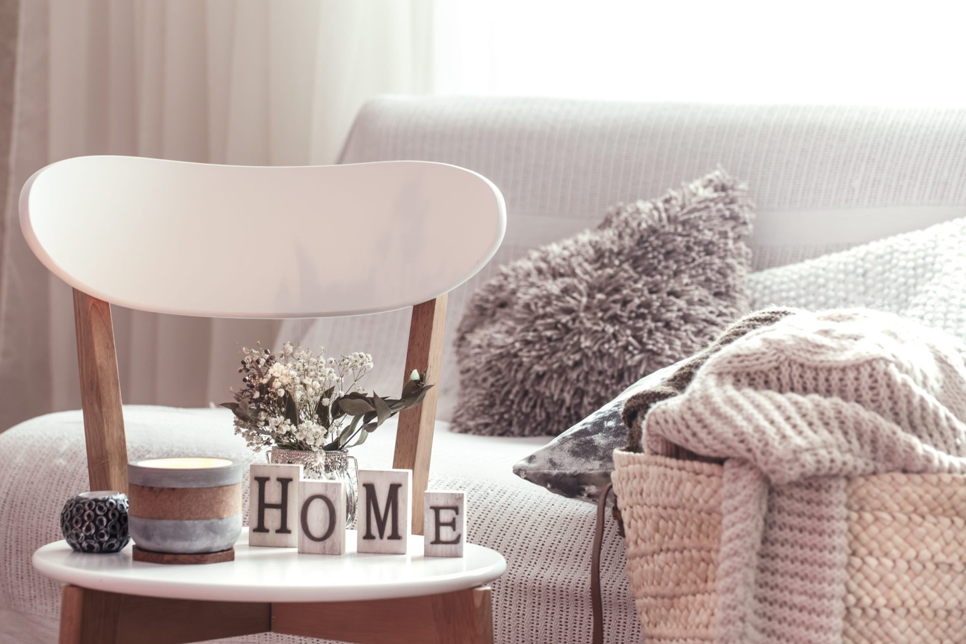 Candles, a vase with flowers with wooden letters of the home on wooden white chair. Sofa and wicker basket with cushions in the background.
